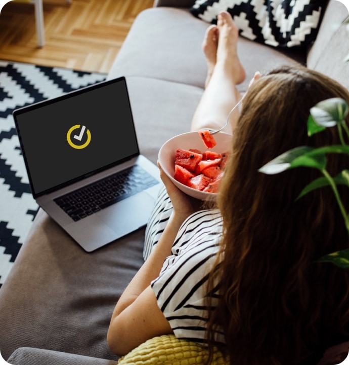 소파에 기대 앉아 그릇에 담긴 과일을 먹는 여성 옆에 Norton 로고가 표시되어 있는 노트북