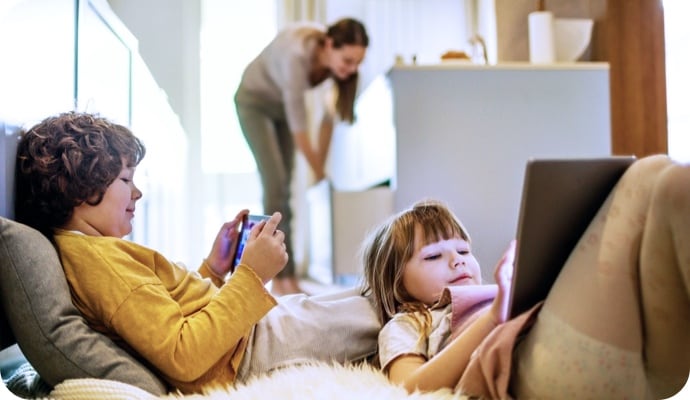 아이들이 누워서 태블릿 장치를 사용하고, 뒤에 있는 어머니는 주방을 정리하는 모습.